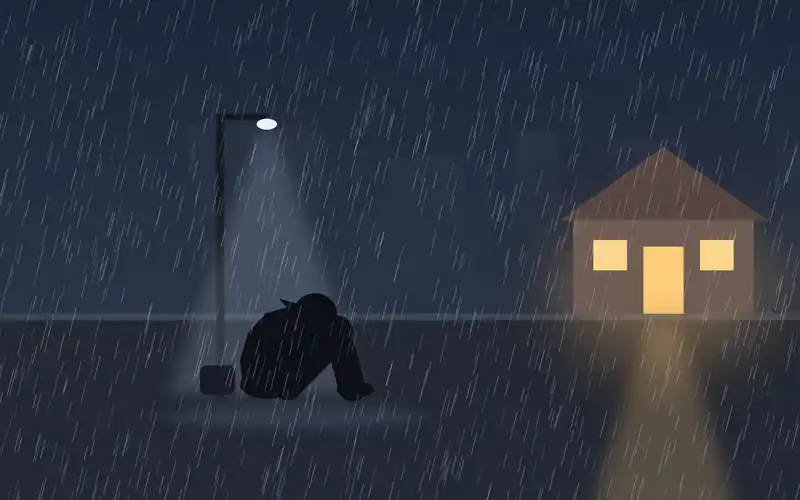 A person sits alone on a wet curb in the pouring rain at night, hood up and head on their knees, while across the street a small house glows with warm light from its open door